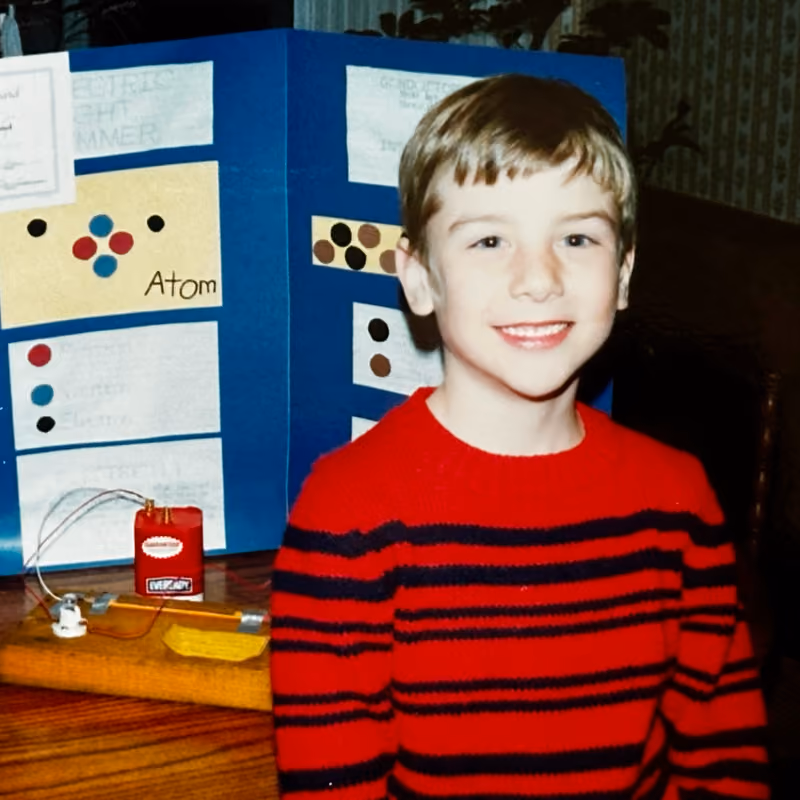 Smiling boy in a red and black striped sweater in front of a science project display board titled Atom.