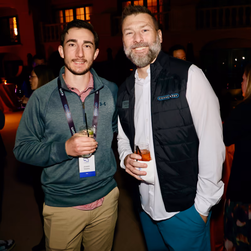 Two men smiling at a social event holding drinks, one wearing a ConnexPay vest.