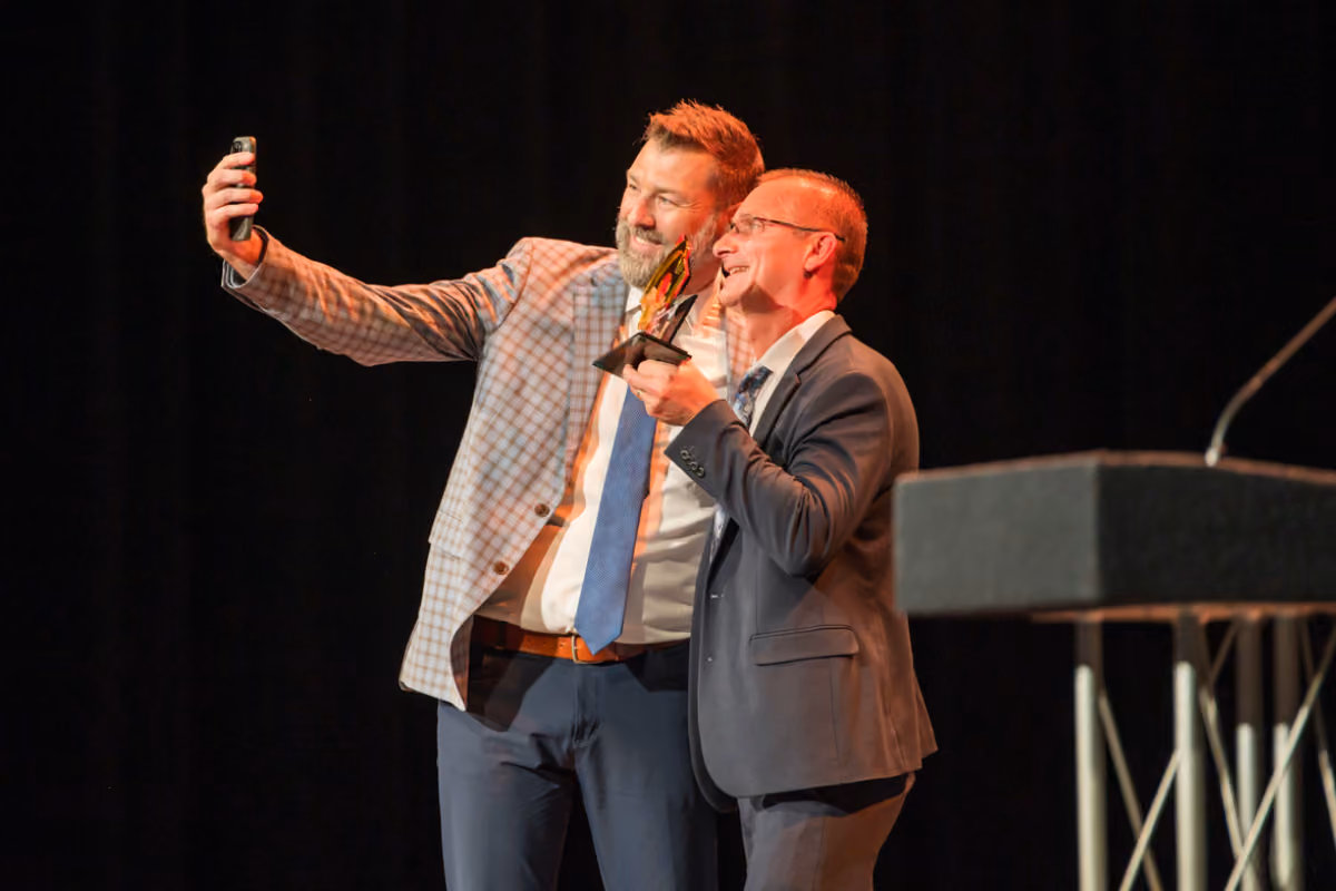Two men in suits smiling and taking a selfie with an award on stage with a dark background.