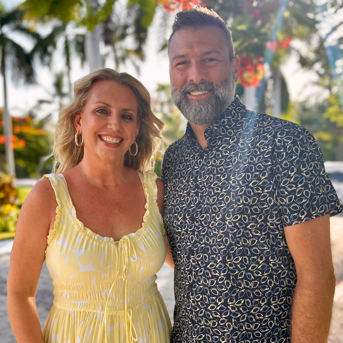 Smiling man with a beard in patterned shirt and woman in yellow dress outdoors with trees in background.