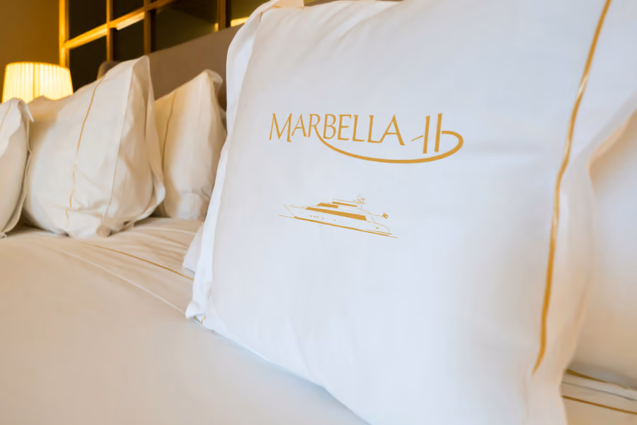 White pillow on a bed with gold embroidery of 'MARBELLA 11' and a yacht illustration.
