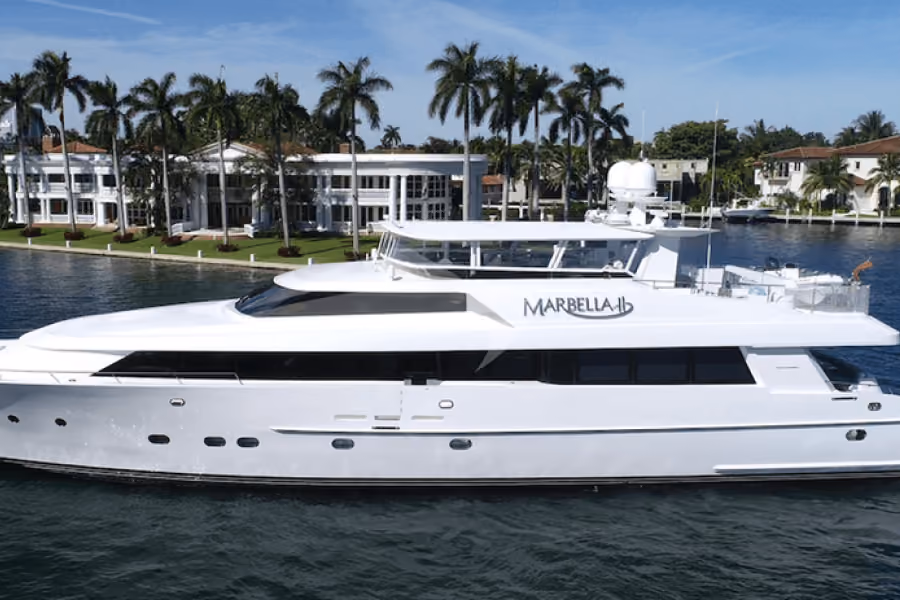 White luxury yacht named Marbella on calm water with palm-lined waterfront homes in background.