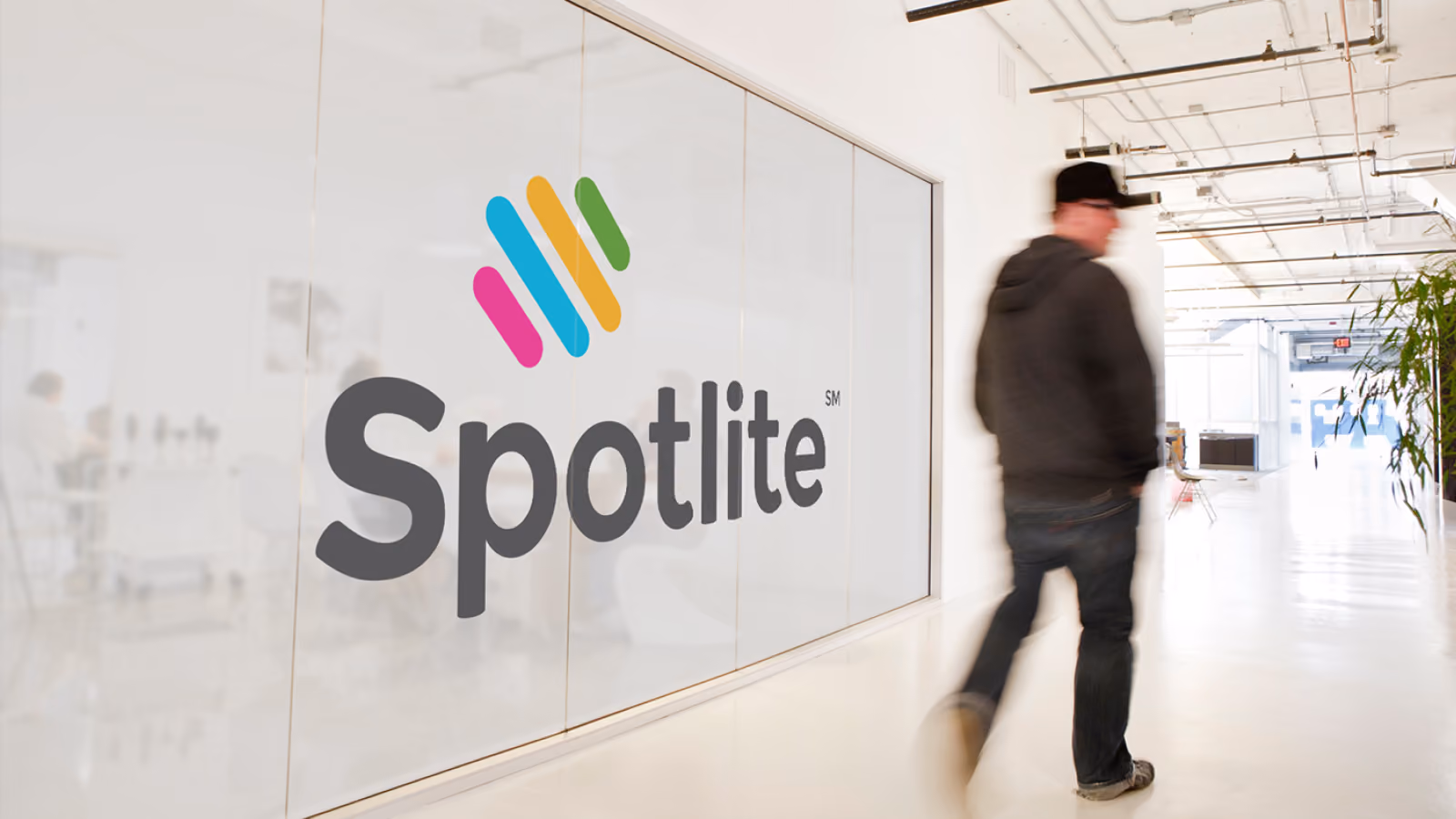 Man in a black cap walking in a bright office hallway past a glass wall with the Spotlite logo.