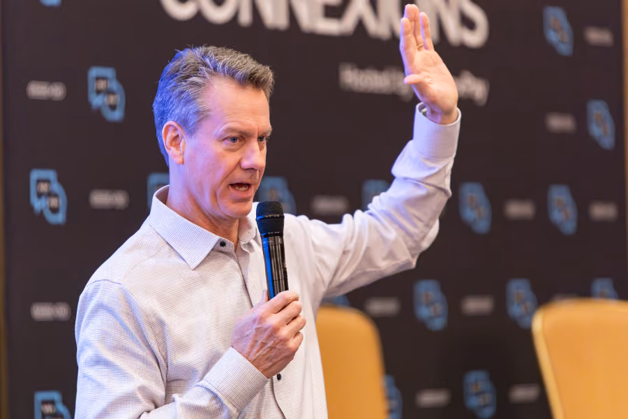 Man in a light shirt speaking into a microphone with one hand raised at a Connexions event.