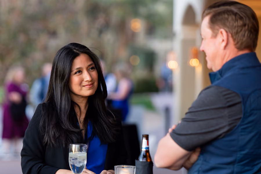 Woman with long dark hair smiling and talking to a man at an outdoor social event with drinks on table.