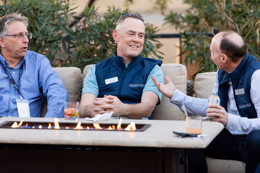 Three men sitting on an outdoor couch talking around a fire table with drinks in hand.