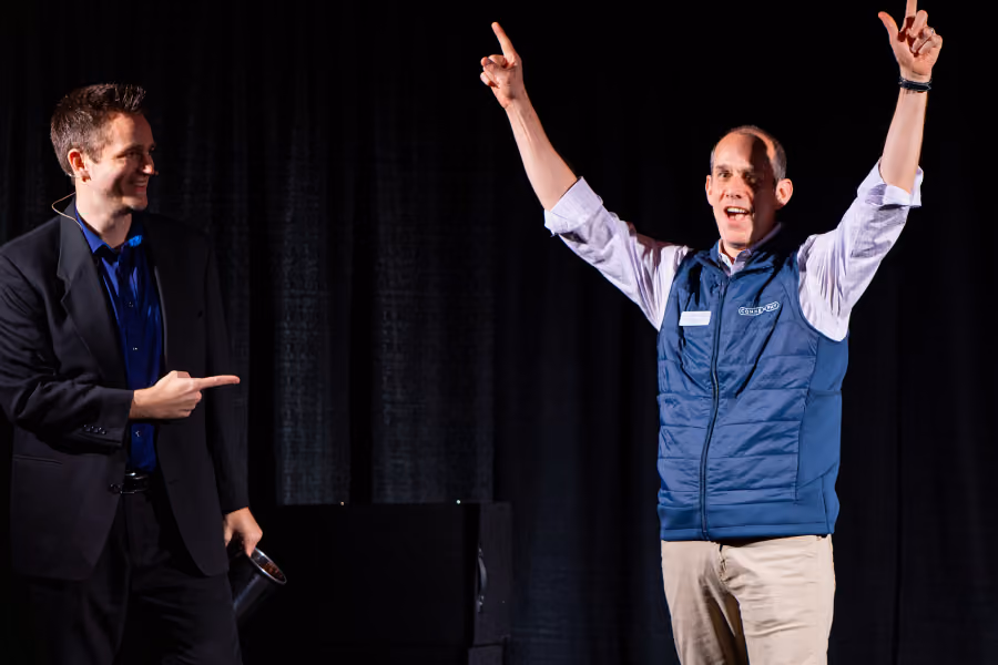 Man in blue vest cheering with hands raised while another man in suit points at him on stage.