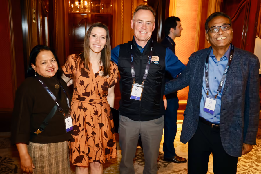 Four smiling people at a conference with badges, standing indoors near wooden walls and carpeted floor.