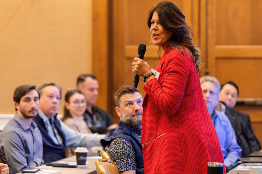 Woman in red jacket speaking into a microphone to attentive seated audience in meeting room.