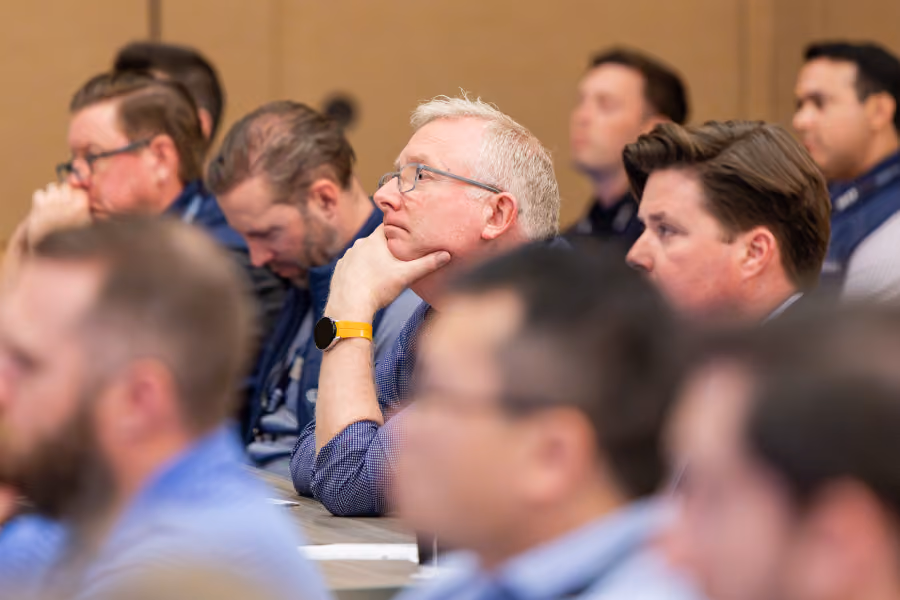 Men attentively listening in a conference or seminar setting, seated in rows.