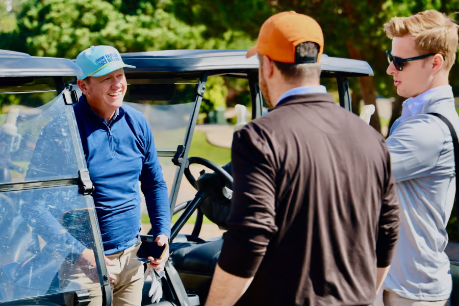 Three men chatting beside a golf cart on a sunny day, one wearing a teal cap and another an orange cap.