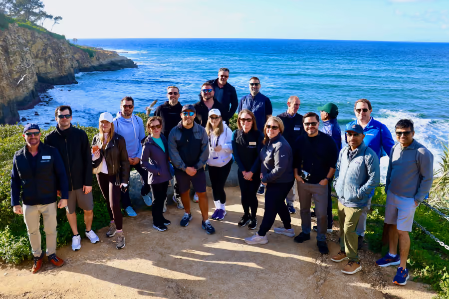 Group of people posing at a coastal viewpoint with cliffs and ocean in the background on a sunny day.