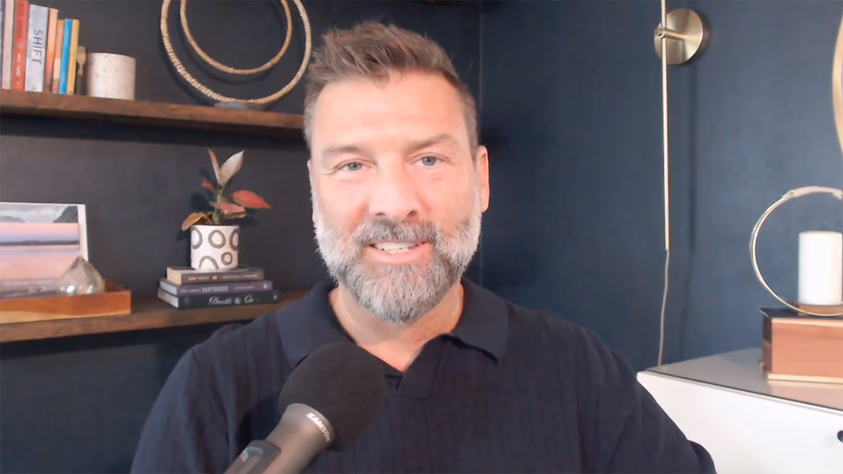Justin speaking into a microphone with dark blue wall and shelves with decor in background.