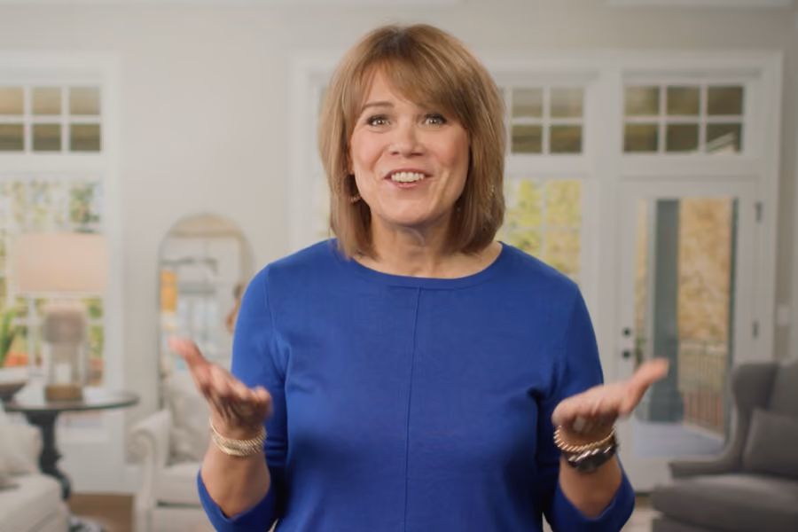 Smiling course leader in a blue sweater gesturing with hands in a bright, cozy living room.