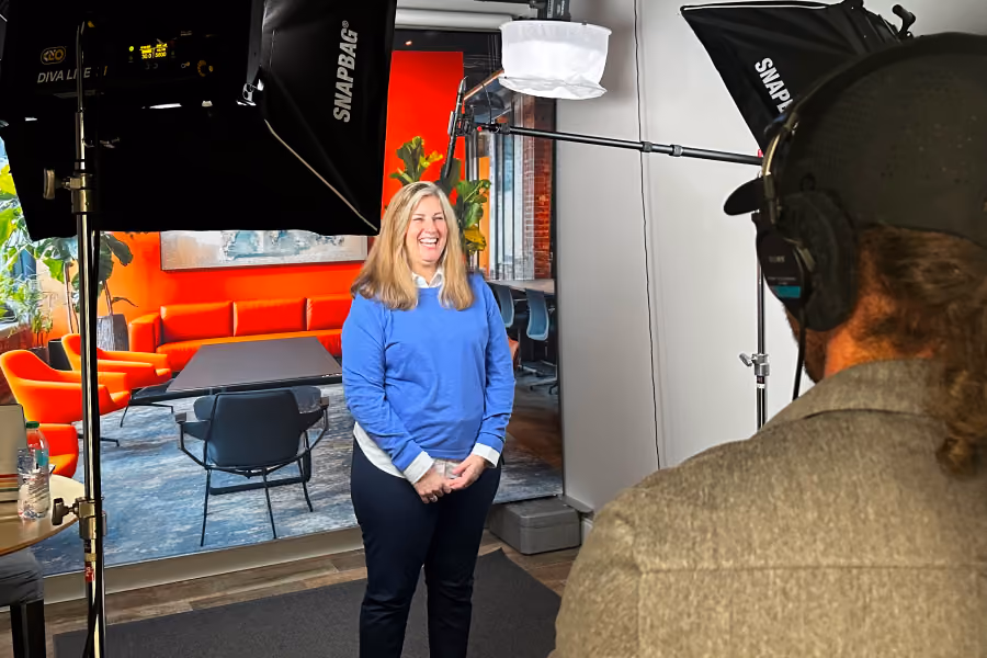 Woman in blue sweater smiling on a brightly colored office set while a man with headphones films.