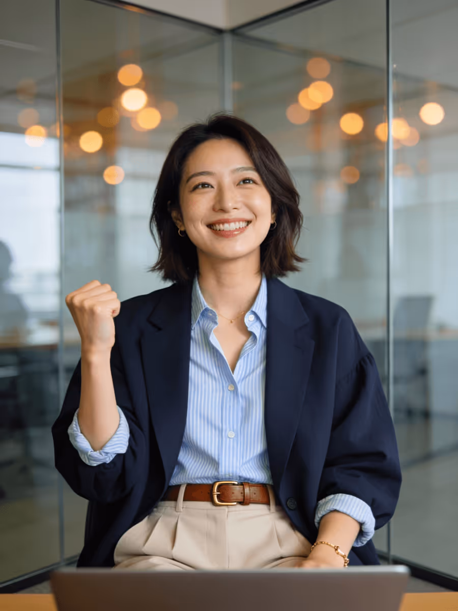Smiling businesswoman in blazer at desk, raising fist in a confident gesture.