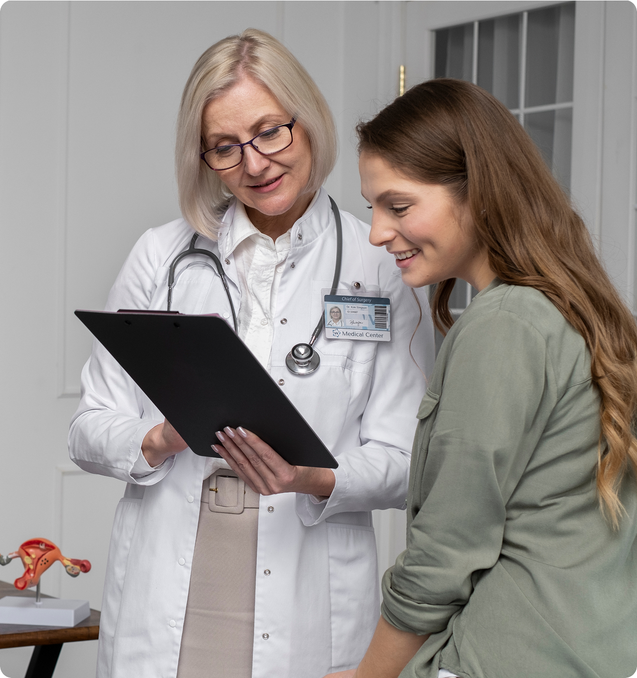 A female doctor in a white coat and glasses reviewing medical charts on a clipboard with a smiling young female patient.