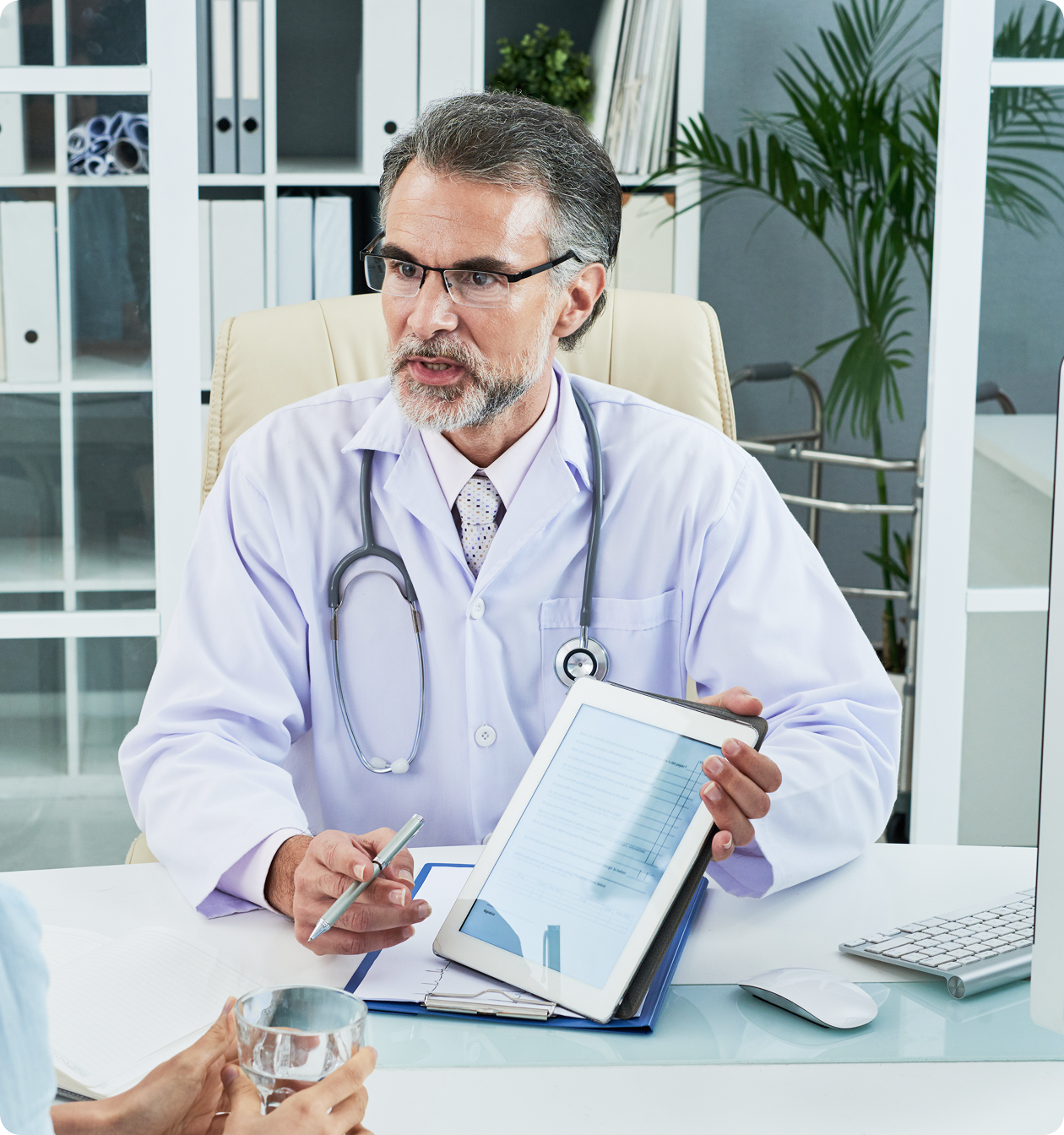 A male doctor with a beard and glasses sitting at a desk and holding out a tablet to a patient (unseen) to show medical data.