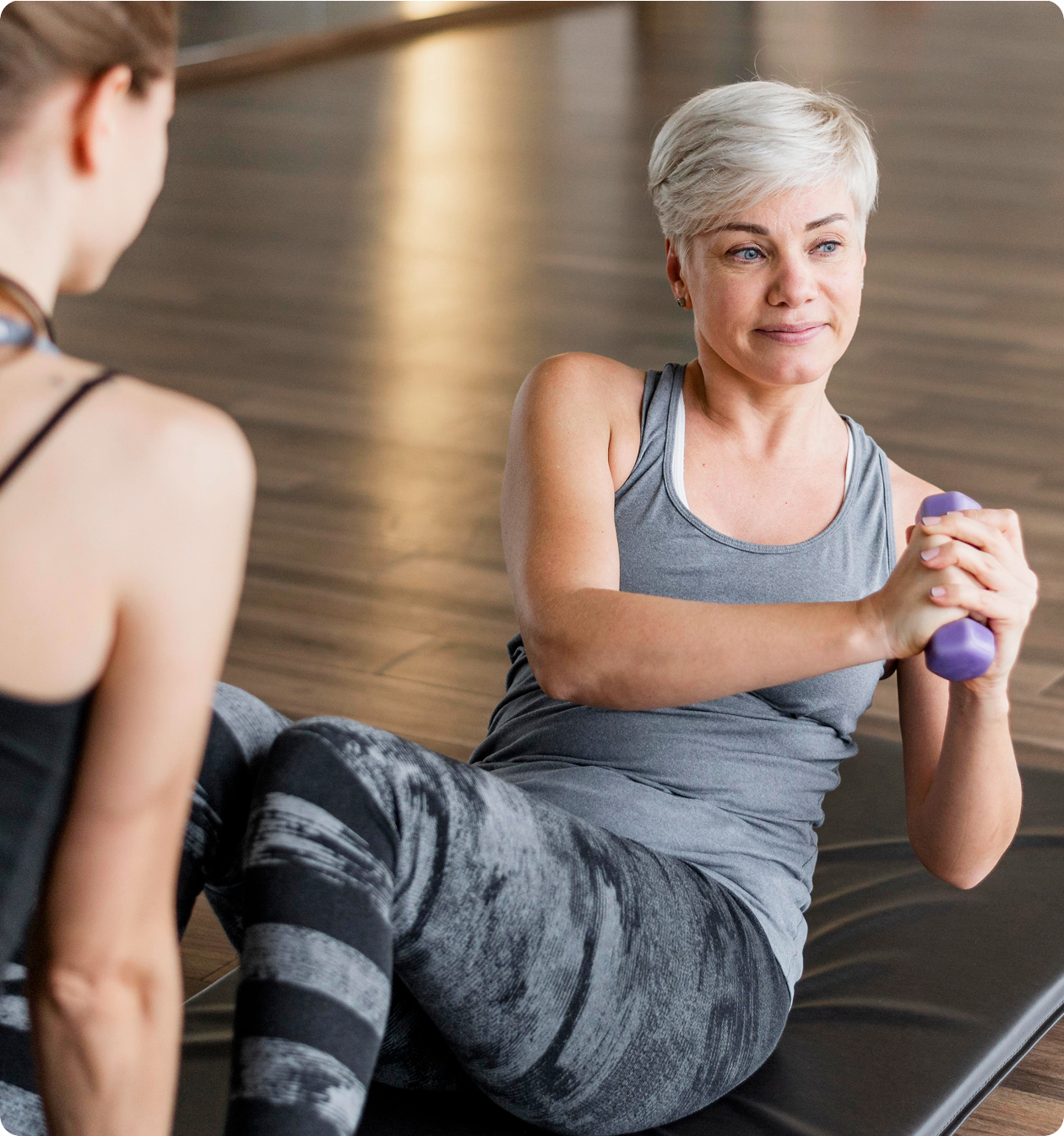 A senior woman with short blonde hair performing core twists with a small purple dumbbell while sitting on an exercise mat, with a trainer visible in the foreground.