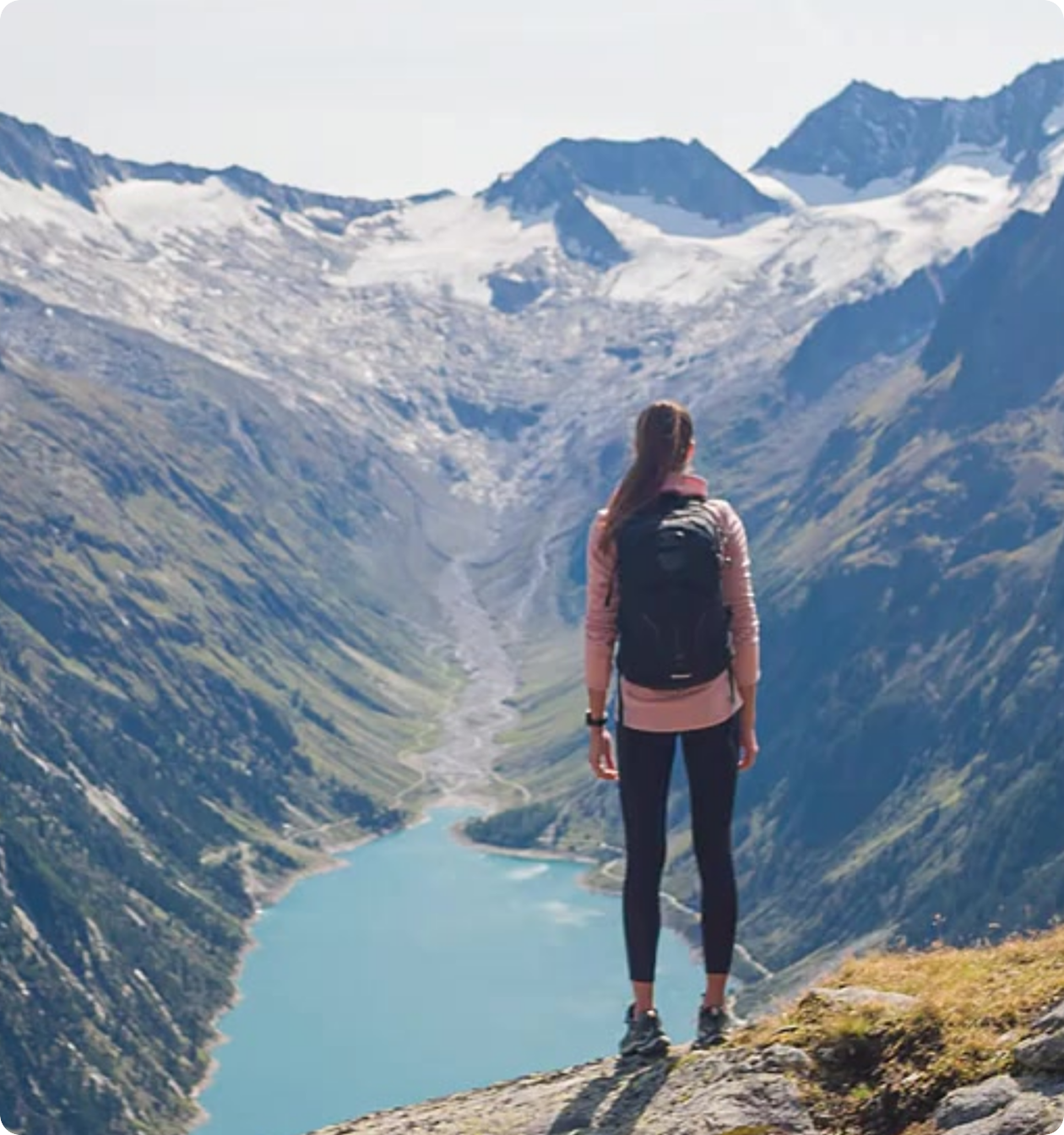 A female hiker with a backpack standing on a peak looking down at a blue mountain lake in a valley, with snow-capped peaks in the background.