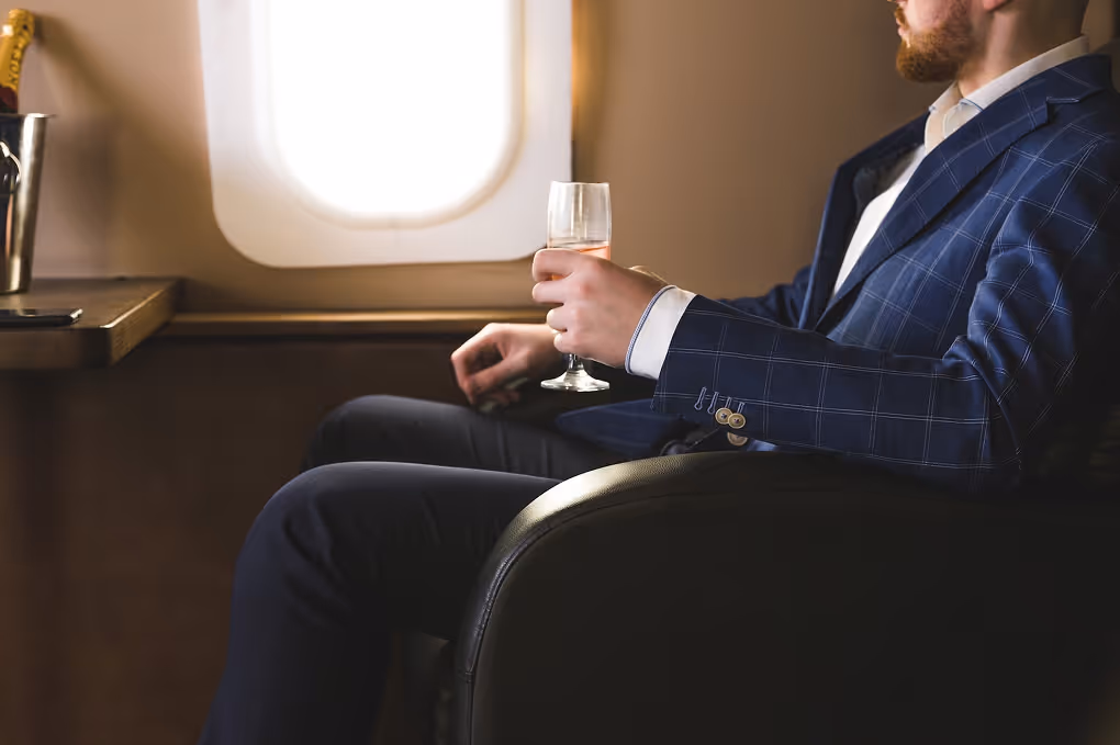 Man in blue suit sitting in airplane seat holding a glass of champagne by the window.