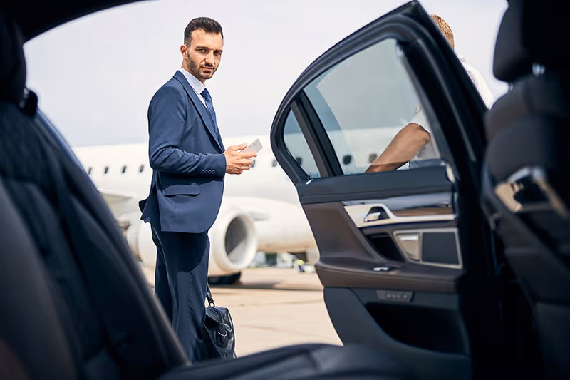 Man in navy blue suit holding phone stands next to open car door with a charter jet in the background.