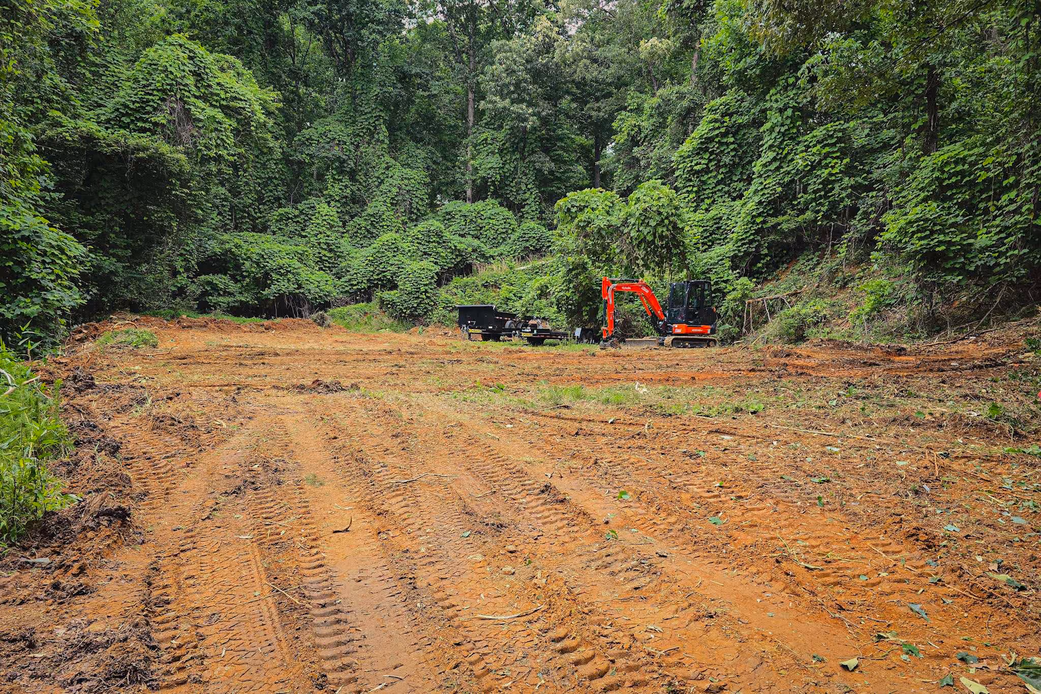 Cleared dirt area with tire tracks and a small red excavator near green forest trees.