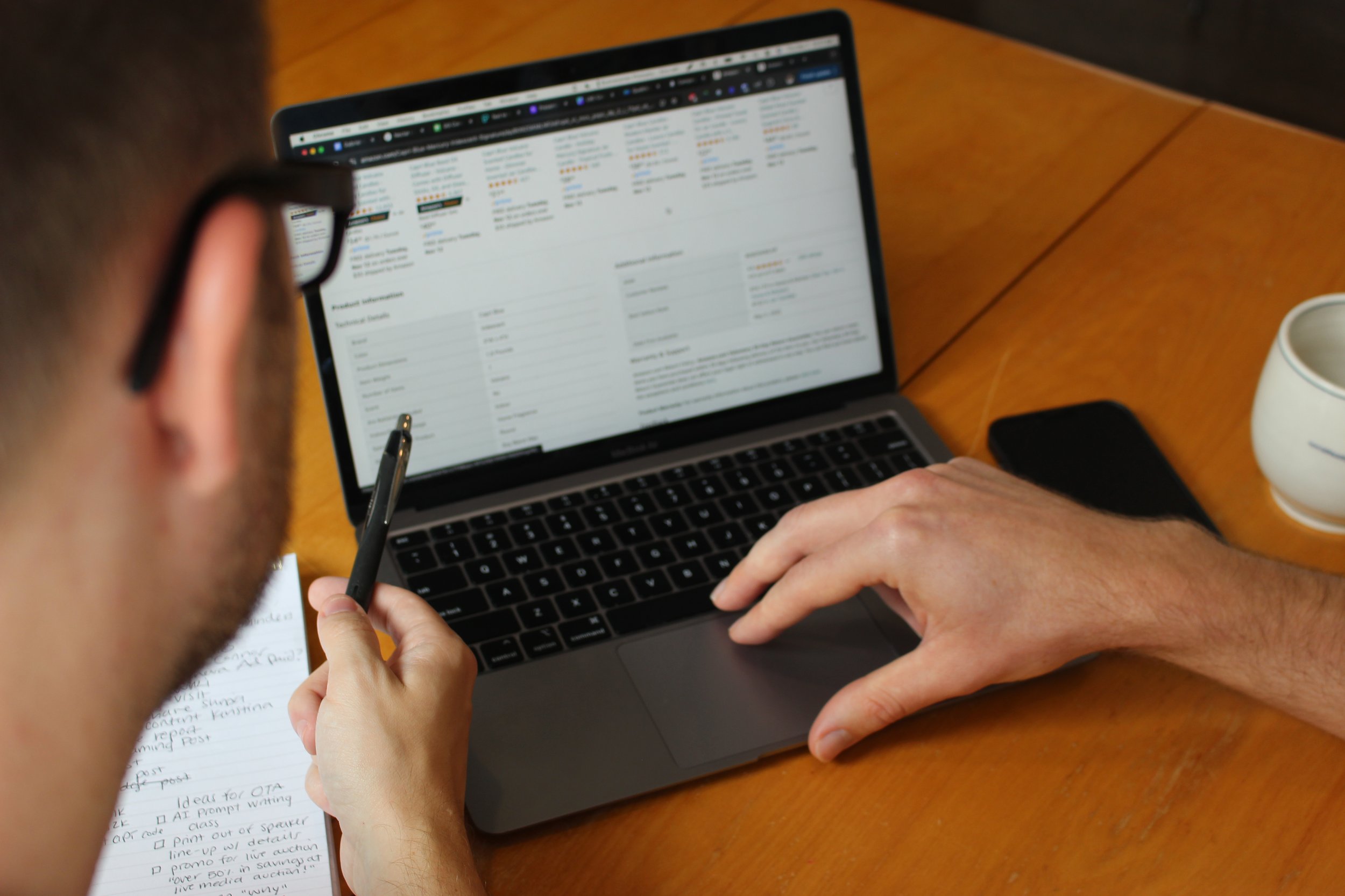 Person holding a pen and using a laptop displaying a detailed product info webpage on a wooden table.