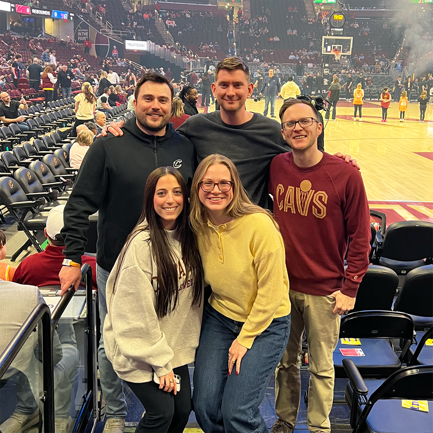 Five people smiling at a basketball arena with the court and spectators behind them.