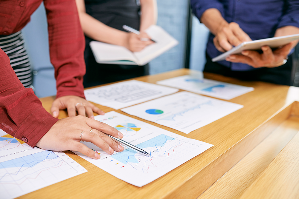Three business people analyzing charts and graphs on paper and a tablet at a wooden table.