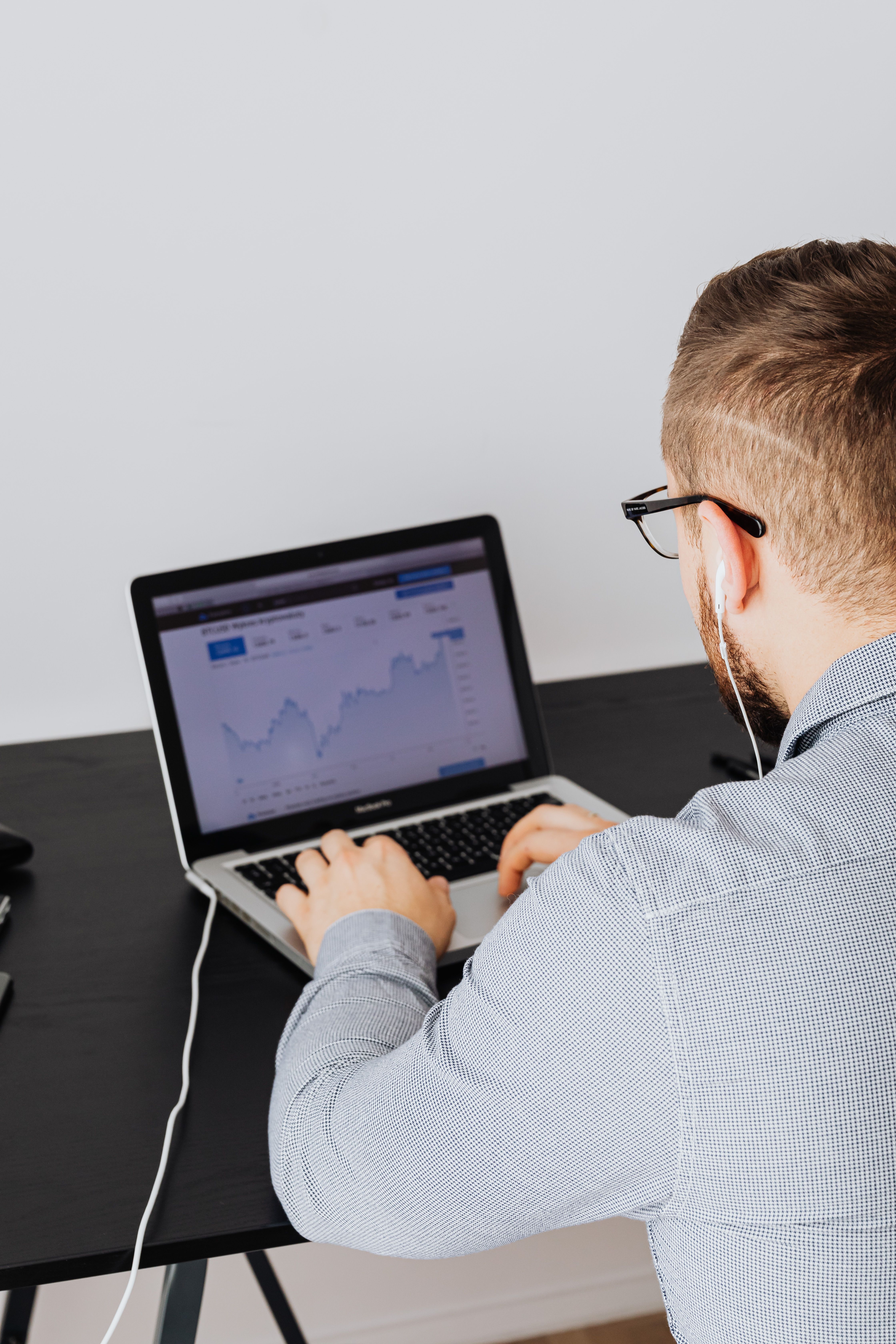 Man wearing glasses and earphones working on a laptop with a graph on the screen.