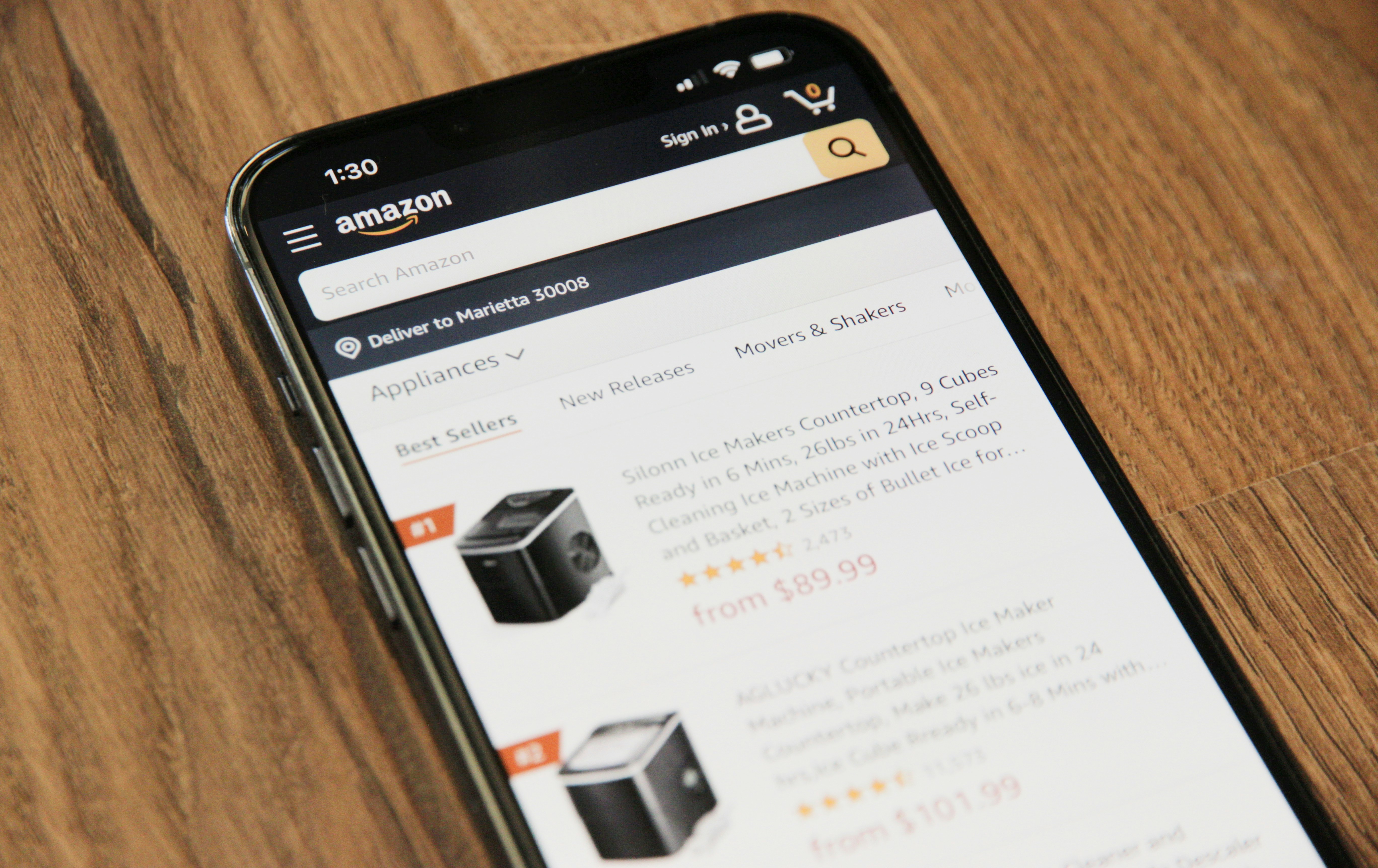 Smartphone showing Amazon app with best-selling ice maker listings on a wooden surface.
