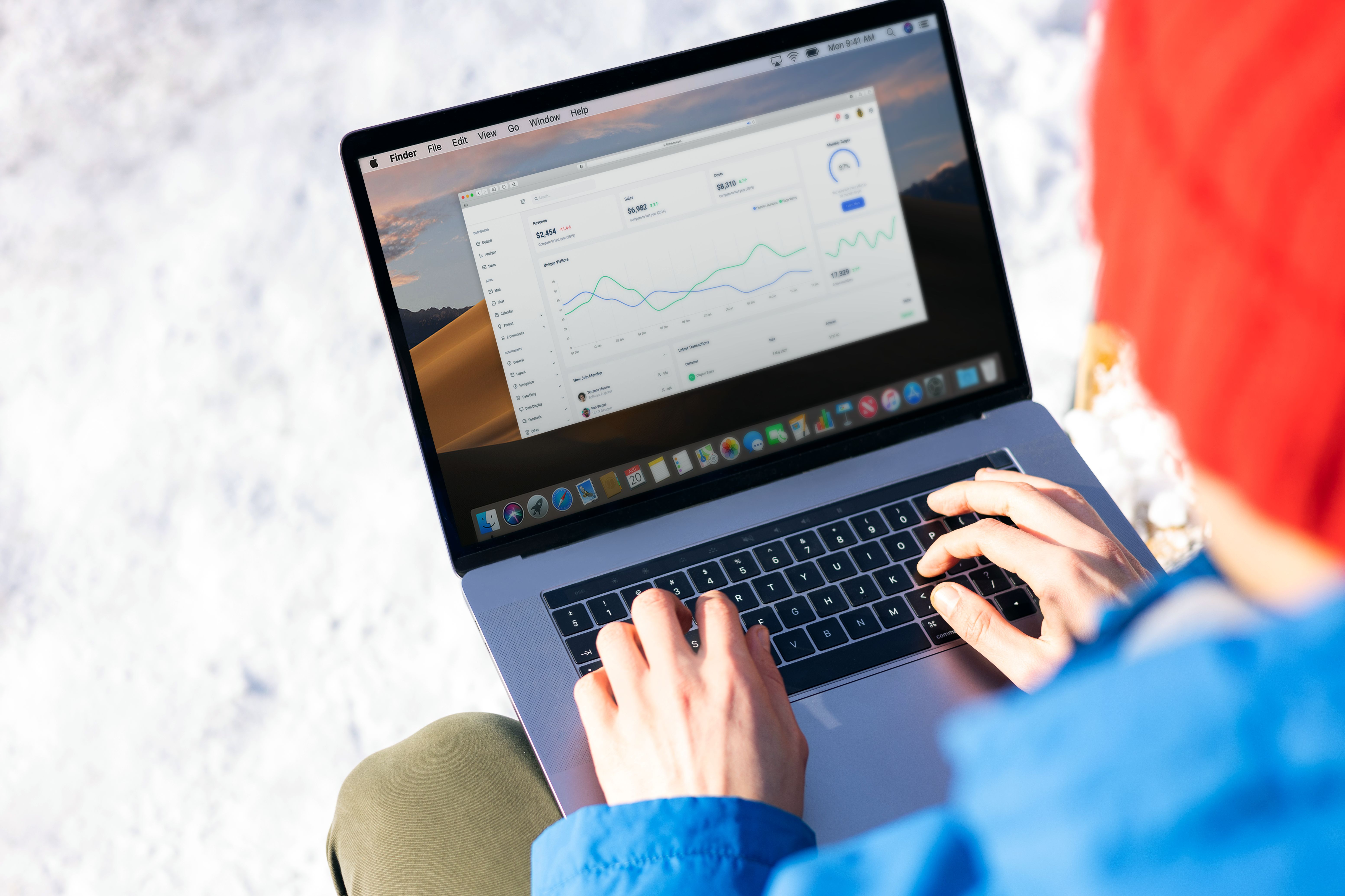 Person wearing red hat and blue jacket typing on laptop with data charts on screen against snowy background.
