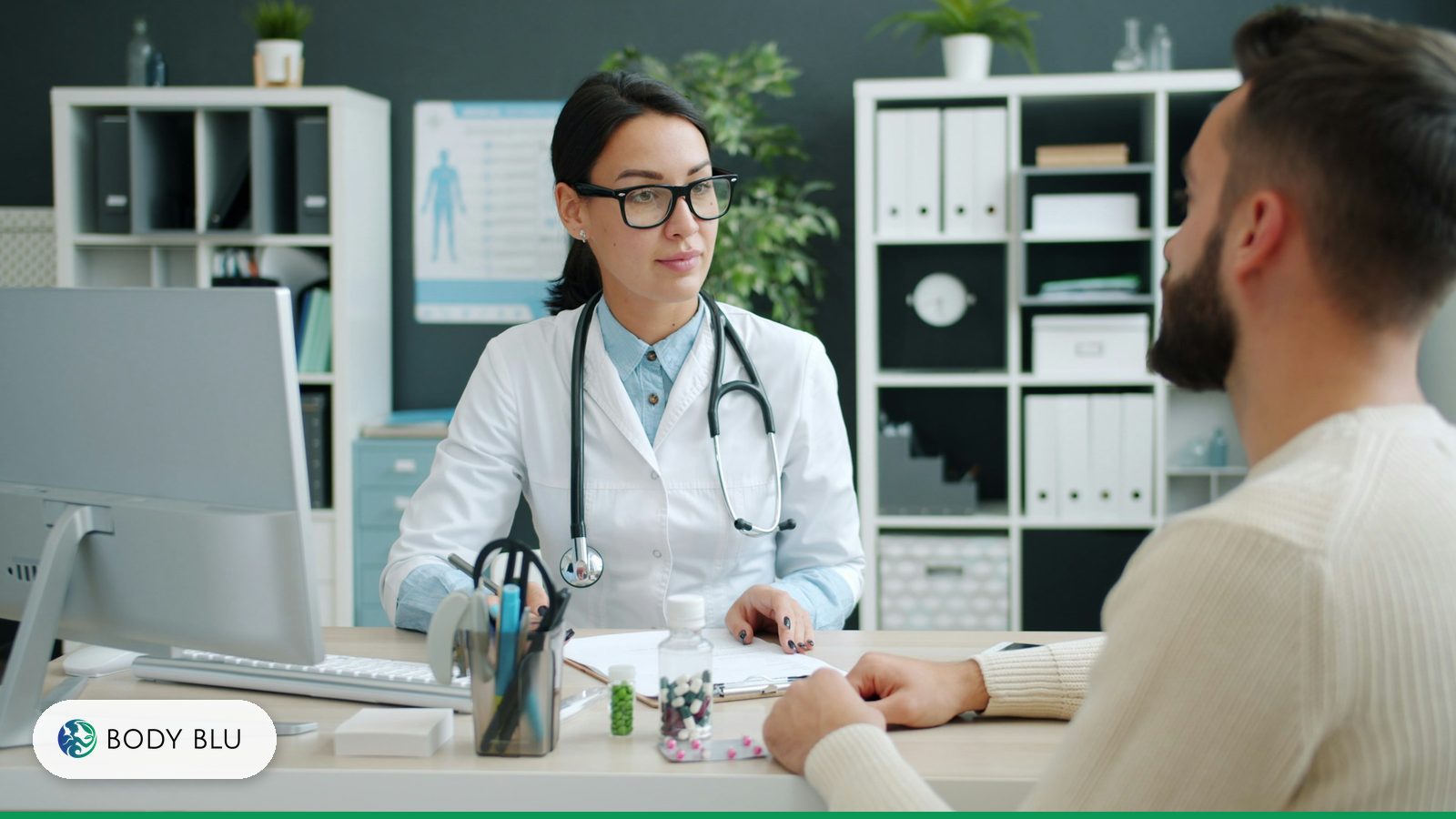 Female doctor with glasses and a stethoscope listening to a male patient across a desk during a clinic consultation