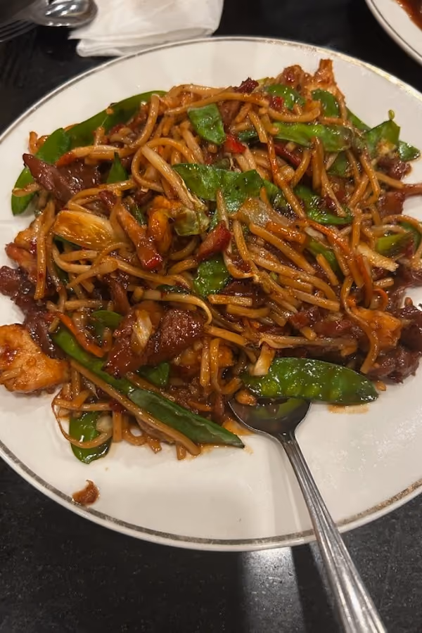 Plate of stir-fried noodles with beef, green snap peas, and vegetables with a spoon on white plate.
