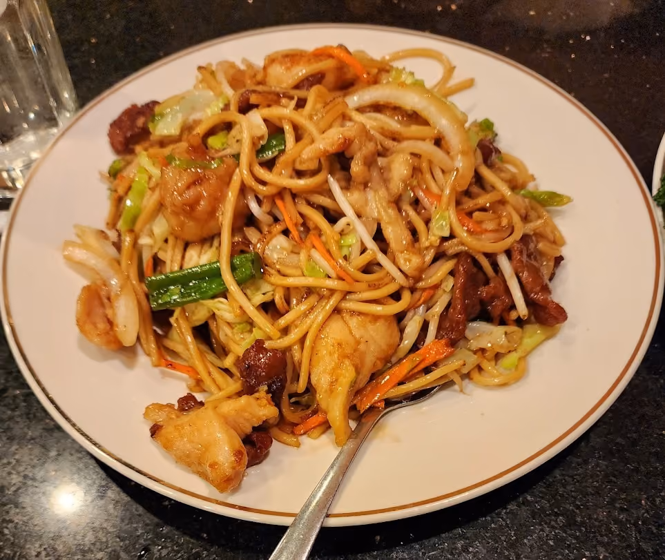 Plate of stir-fried noodles with chicken, vegetables, and a spoon on a dark table.