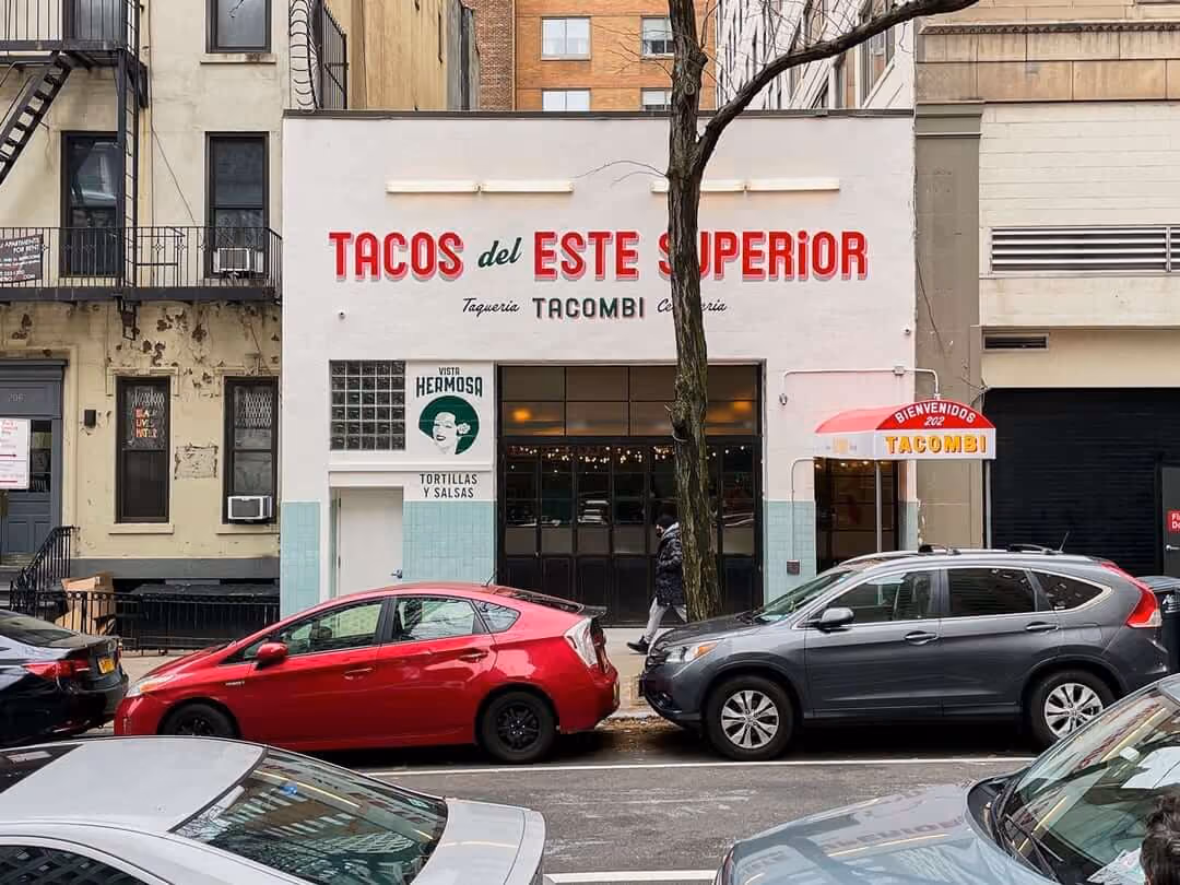Street view of a Tacombi restaurant storefront with a painted 'Tacos del Este Superior' sign, parked cars lining the curb