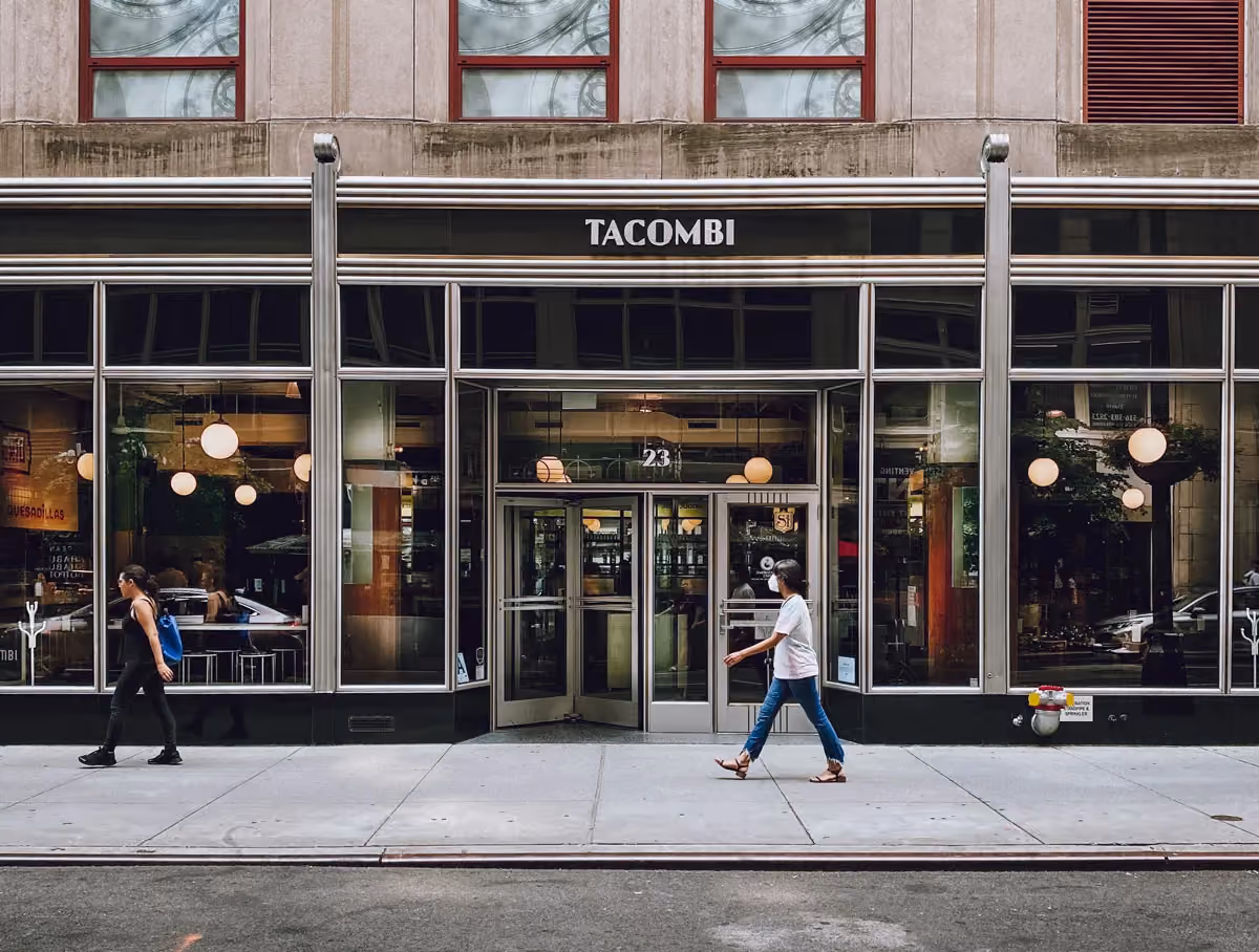 Sleek glass storefront of Tacombi with metal framing and revolving door, pedestrians walking on the sidewalk out front