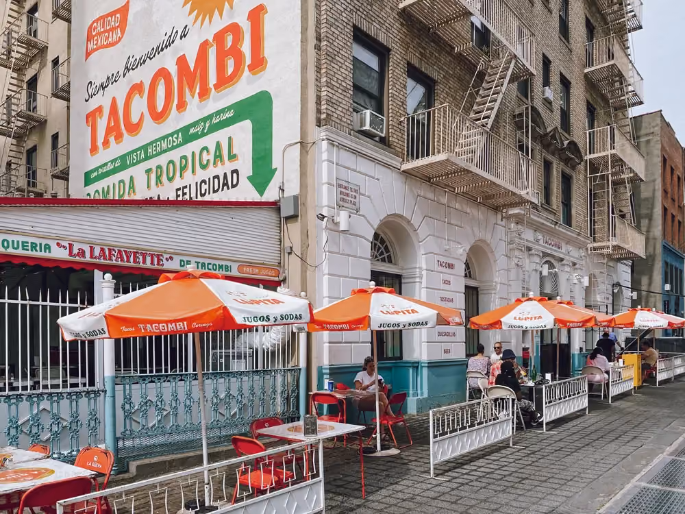 Tacombi taqueria storefront with a large painted mural sign and outdoor seating under orange umbrellas, patrons dining at sidewalk tables