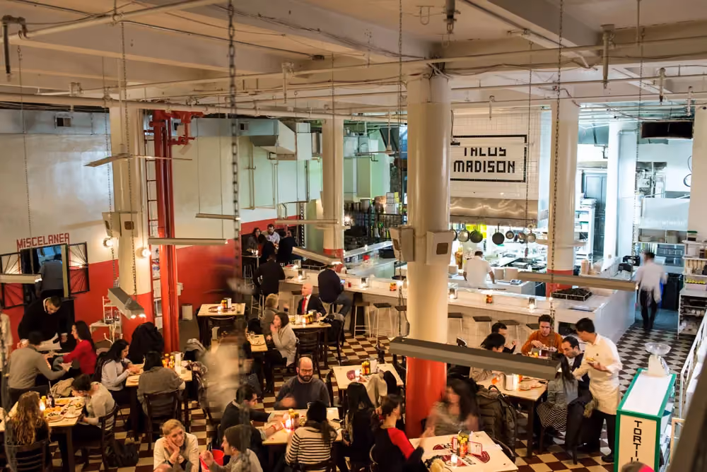 Overhead view of a bustling restaurant with checkered floors, red columns, and diners seated at multiple tables near an open kitchen