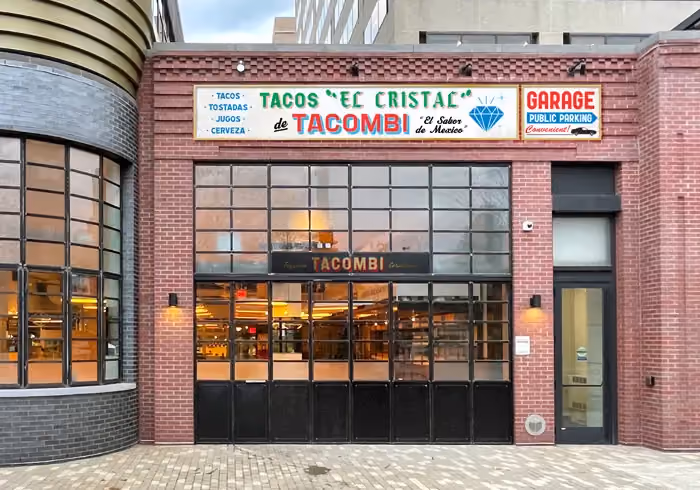 Brick storefront of 'Tacos El Cristal de Tacombi' with large paned windows and a garage parking sign beside it