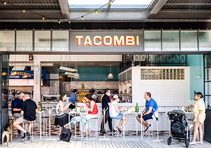 Outdoor patio of Tacombi restaurant with people seated at high-top tables under an illuminated sign