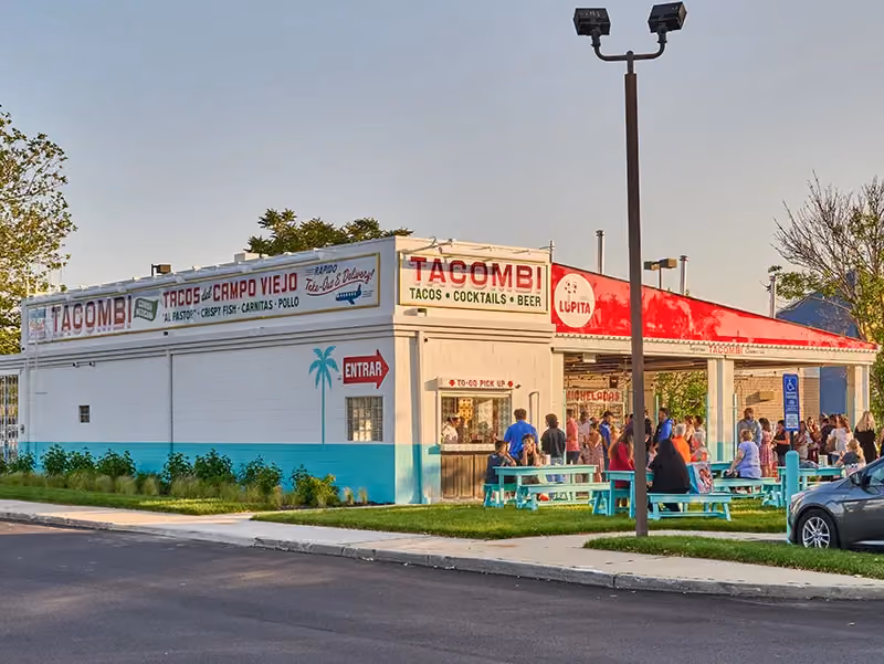 Standalone Tacombi restaurant building with a red awning, outdoor picnic tables, and people gathered outside at dusk