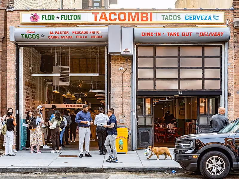 Brick storefront of 'Flor de Fulton Tacombi' with a line of customers waiting outside and a dog walking on the sidewalk