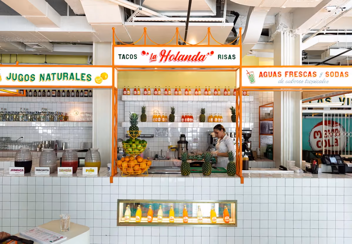 Colorful juice and aguas frescas stand called 'La Holanda' with fresh pineapples, limes, and oranges on display, an employee preparing drinks behind the counter.