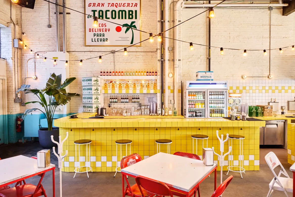 Interior of a Tacombi restaurant with a bright yellow tiled bar, string lights, and red dining tables