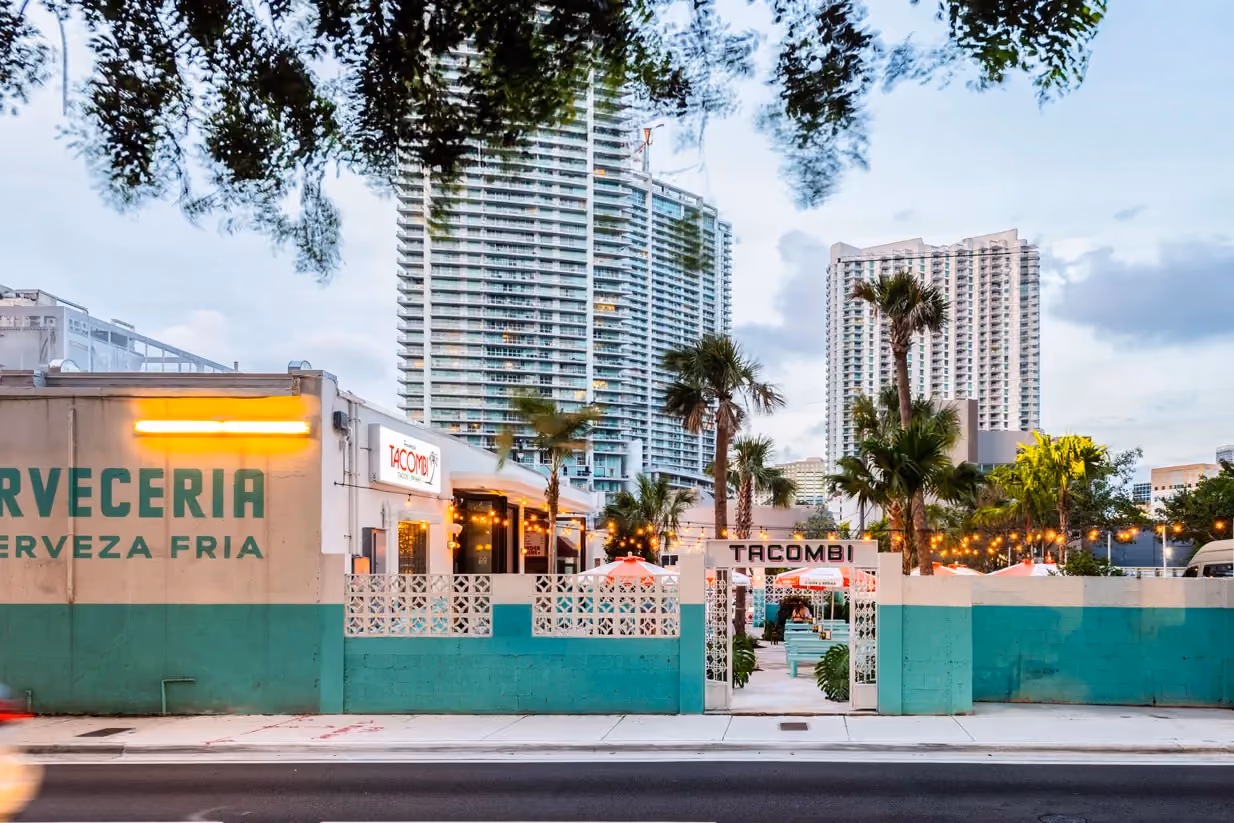 Tacombi restaurant entrance with a teal courtyard wall, palm trees, and string lights, city high-rises visible in the background at dusk