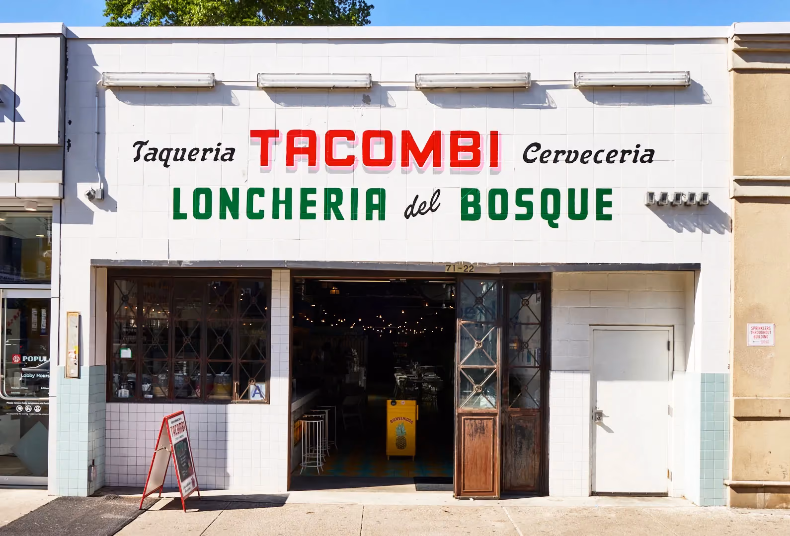 White tiled storefront reading 'Taqueria Tacombi Cerveceria, Loncheria del Bosque' with an open entrance and A-frame sidewalk sign