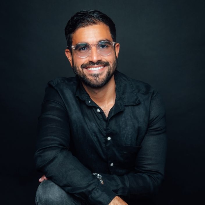 Homme souriant avec lunettes transparentes et chemise noire, assis devant un fond noir.