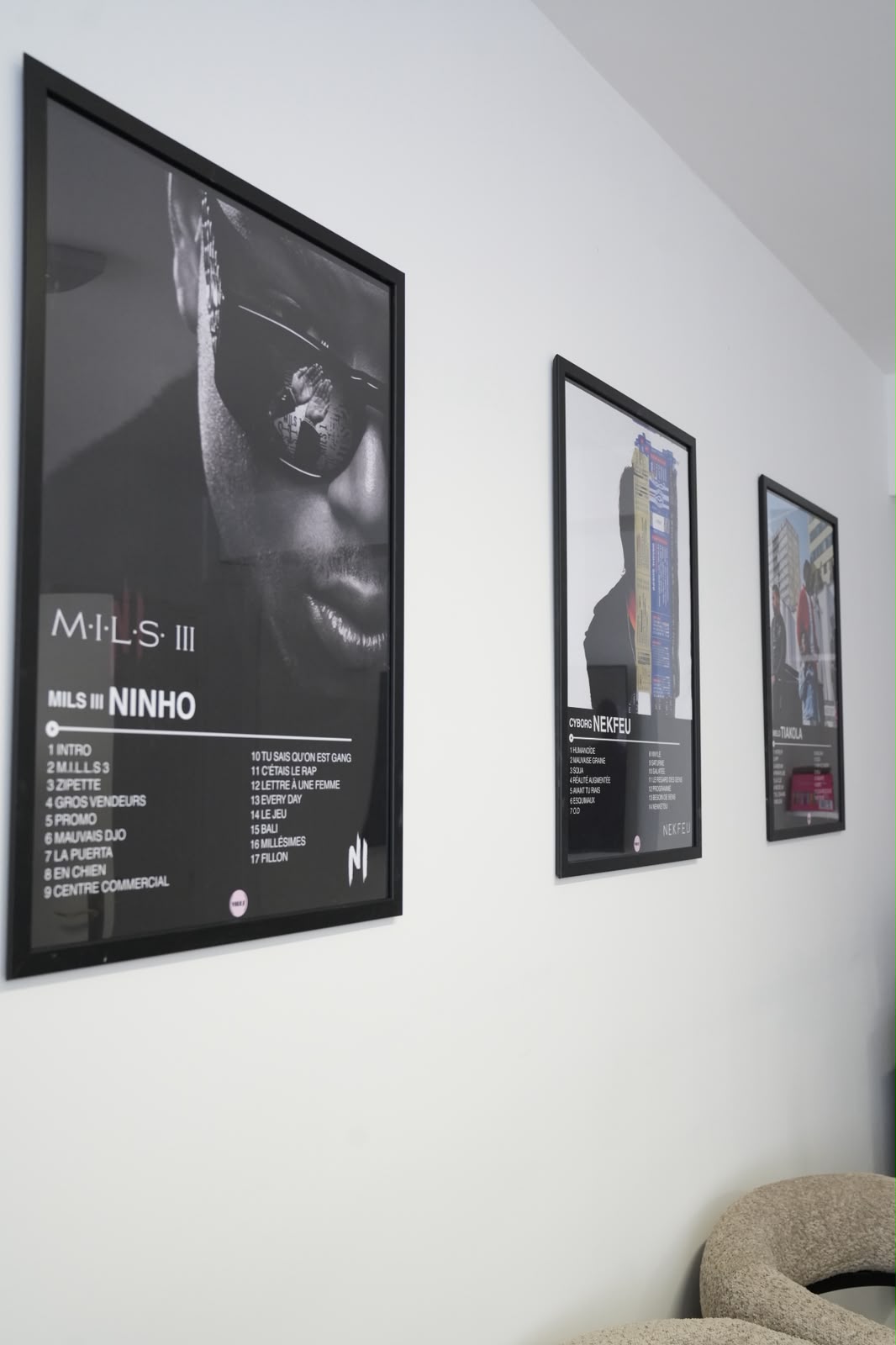 Trois affiches encadrées sur un mur blanc avec une chaise beige en bas à droite.